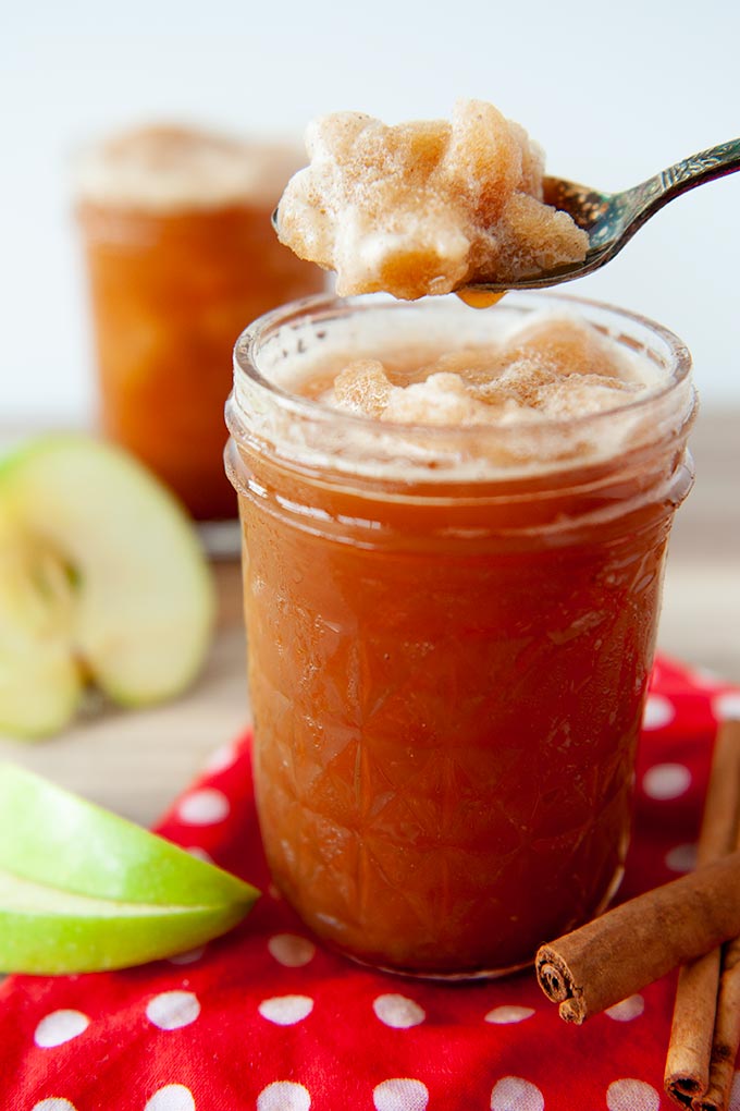 Up close apple cider slushie in a glass with a spoonful of slushie above it, cut up apples and cinnamon sticks