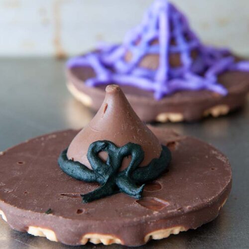 Two cookie witch hats, one decorated in frosting as a purple spider web and the other has a black bow