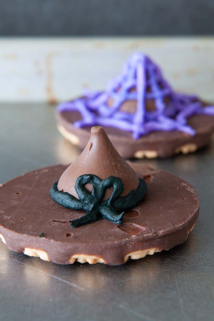 Two cookie witch hats, one decorated  in frosting as a purple spider web and the other has a black bow 