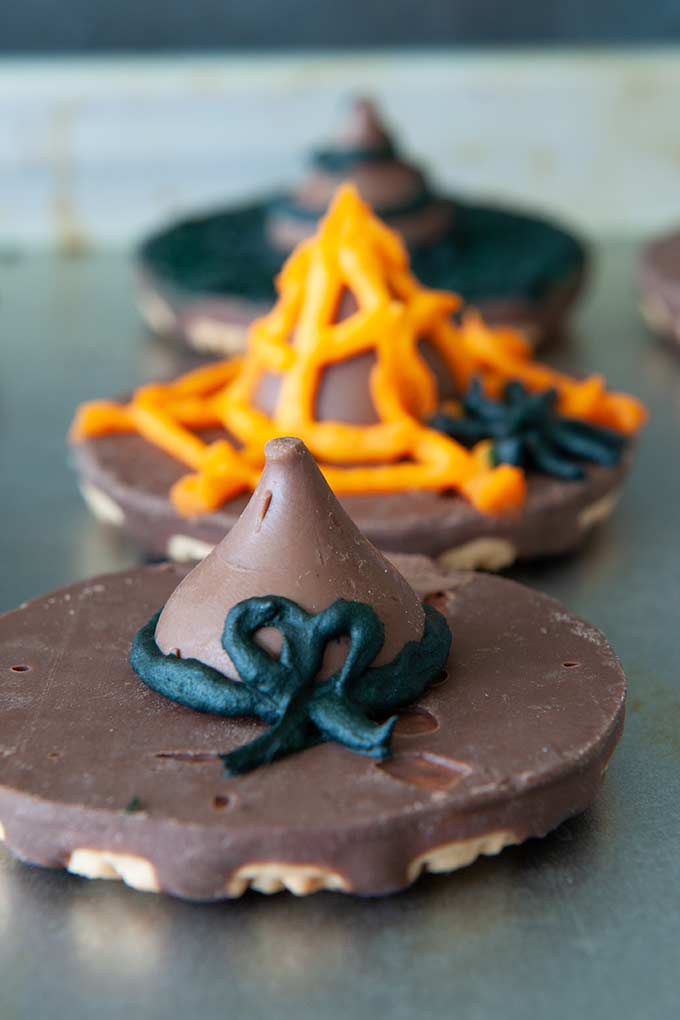 Three cookie witch hats, decorated in frosting in orange spider web with a small black spider, a black swirl and the last one has a black bow 