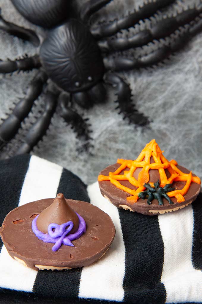 Two cookie witch hats, decorated in frosting one with purple bow and the other with orange spider web with a big black spider in the background