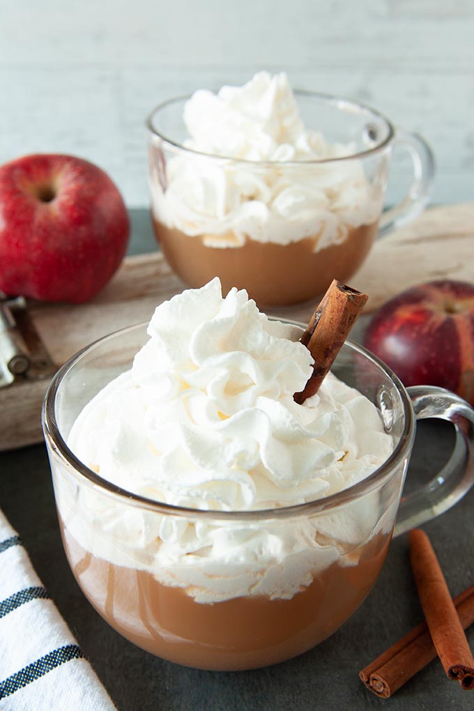 Two clear mugs of apple cider cocktail topped with whipped cream and a cinnamon stick
