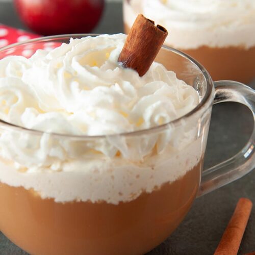 Two clear mugs of apple cider cocktail topped with whipped cream and a cinnamon stick