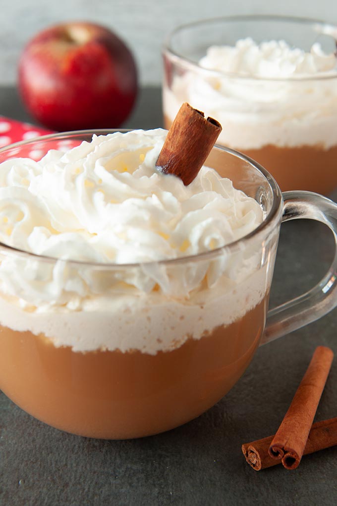 Two mugs of apple cider cocktail topped with whipped cream and a cinnamon stick