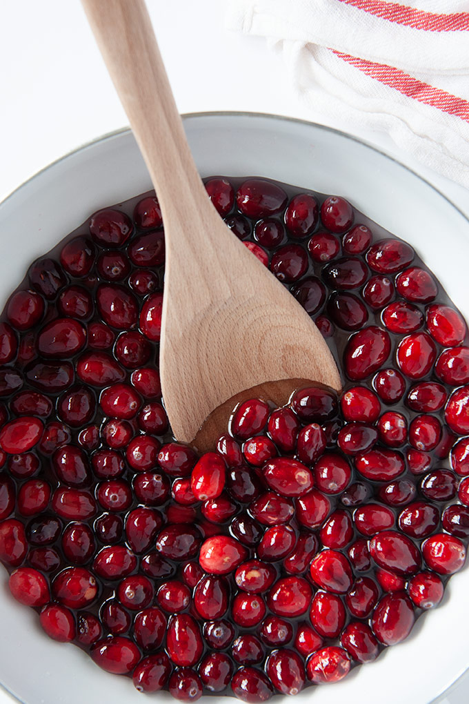 Cranberries cooking in a saucepan of sugar syrup with a wooden spoon.