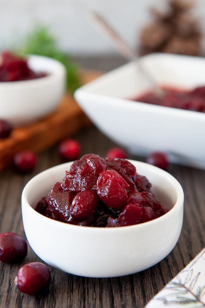 A small bowl full of cranberry sauce, fresh cranberries scattered around, and a square serving dish in the back.