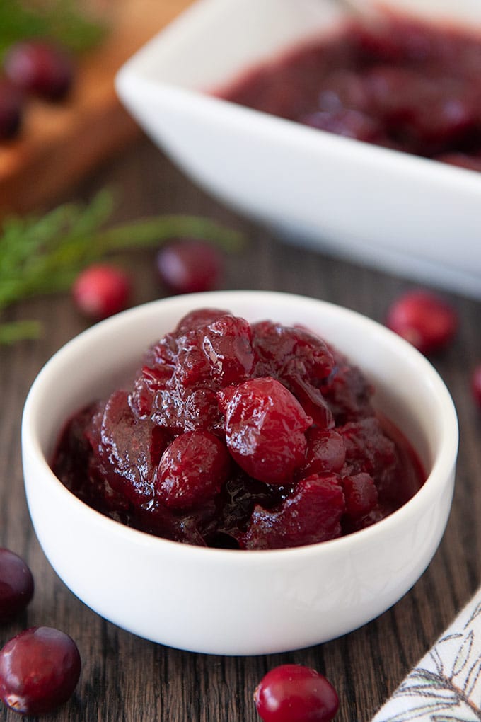 A small bowl full of cranberry sauce, fresh cranberries scattered around, and a square serving dish in the back.