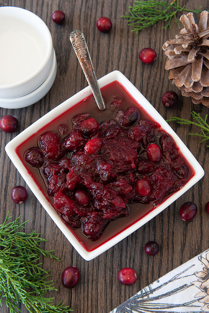 A spoon in a square serving dish of fresh cranberry sauce with cranberries sprinkled around, a pinecone, and some greenery.
