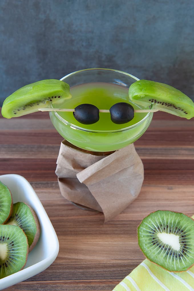 A coupe glass filled with green kiwi martini, slice kiwis for ears and black olives on a cocktail stirrer for eyes.  The glass stem is wrapped in a brown paper bag "coat".  Cut kiwis are in the foreground with a bright yellow towel.