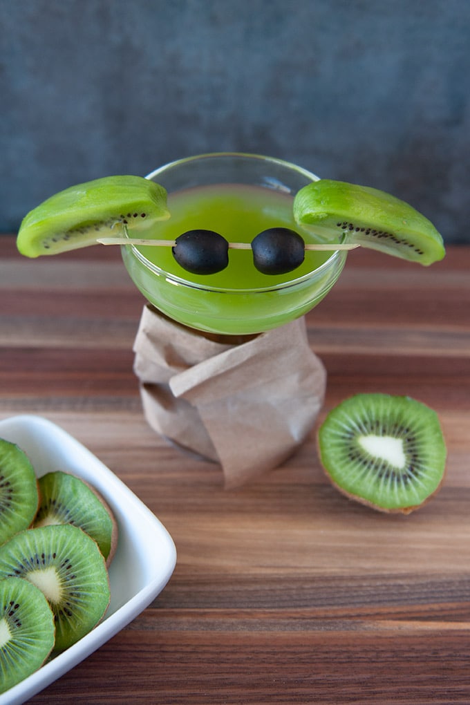 A coupe glass filled with green kiwi martini, slice kiwis for ears and black olives on a cocktail stirrer for eyes.  The glass stem is wrapped in a brown paper bag "coat".  Cut kiwis are in the foreground.