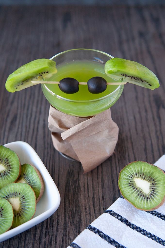 A coupe glass filled with green kiwi martini, slice kiwis for ears and black olives on a cocktail stirrer for eyes.  The glass stem is wrapped in a brown paper bag "coat".  Cut kiwis are in the foreground with a black and white striped towel.