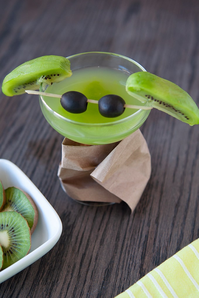 A coupe glass filled with green kiwi martini, slice kiwis for ears and black olives on a cocktail stirrer for eyes.  The glass stem is wrapped in a brown paper bag "coat".  Cut kiwis are in the foreground with a bright yellow striped towel.