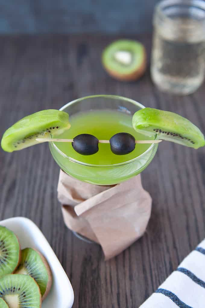 A coupe glass filled with green kiwi martini, slice kiwis for ears and black olives on a cocktail stirrer for eyes.  The glass stem is wrapped in a brown paper bag "coat".  Cut kiwis are in the foreground. with a black and white striped towel.  Glass of simple syrup and cut kiwi in background.