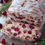 Baked and glazed cranberry bread from above with one slice cut and laying in front, orange slices and cranberries scattered around.