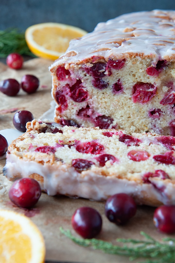 Baked and glazed cranberry bread with one slice cut and laying in front, orange slices and cranberries scattered around.