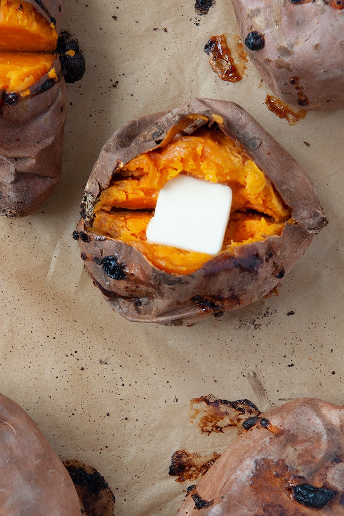 Sweet potato cut open and topped with margarine on brown parchment paper surrounded by a couple other potatoes 