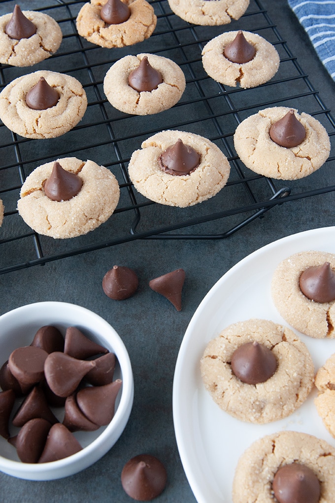 A plate of peanut blossom cookies, a cooling rack of cookies, and a little white bowl of unwrapped chocolate kisses