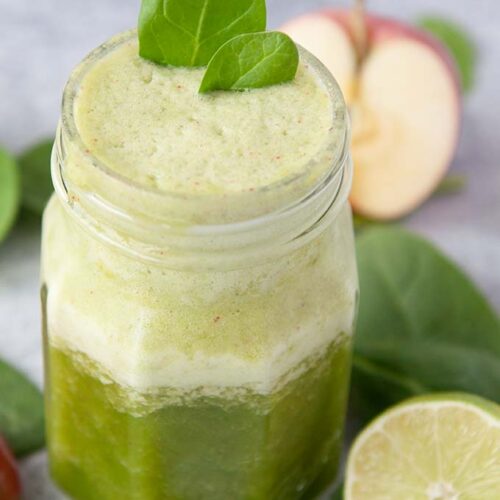 Up close of a jar of green smoothie, with two spinach leaves on top. Cut lime, but apple, and spinach leaves around the jar.