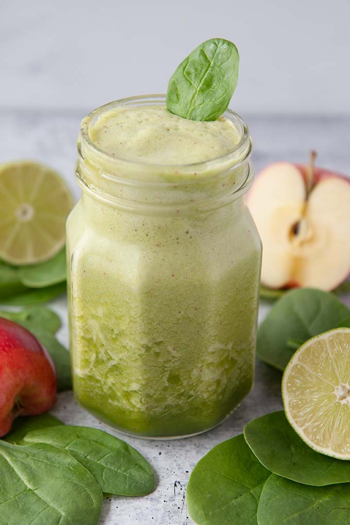 Up close of a jar of green smoothie, with one spinach leaves on top.  Cut lime, but apple, and spinach leaves around the jar.