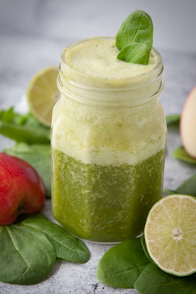 Up close of a jar of green smoothie with two spinach leaves on top.  Cut lime, but apple, and spinach leaves around the jar.