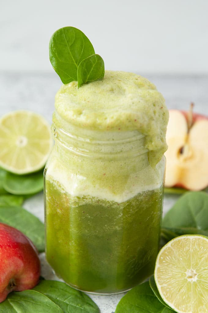 Up close of a jar of green smoothie with overflowing foam and two spinach leaves on top.  Cut lime, but apple, and spinach leaves around the jar.