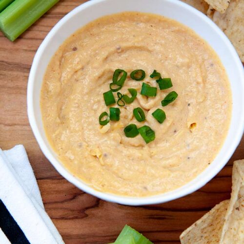 Above view of small white bowl of cheese dip sprinkled with green onions. Chips and celery and white towel with a black stripe.