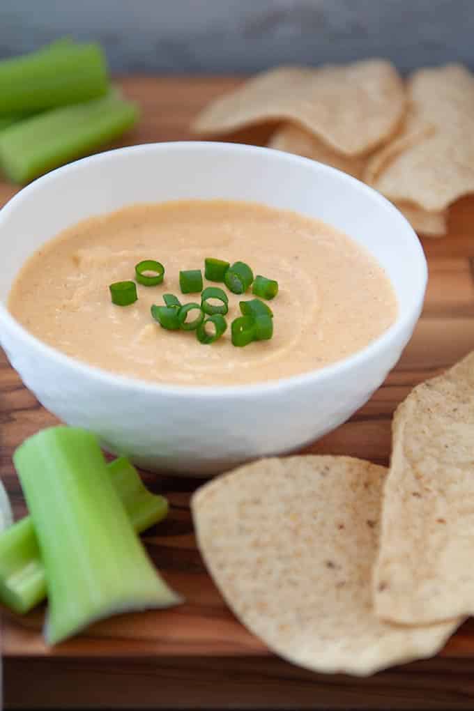 Small white bowl of cheese dip sprinkled with green onions.  Chips and celery in front and back of bowl.