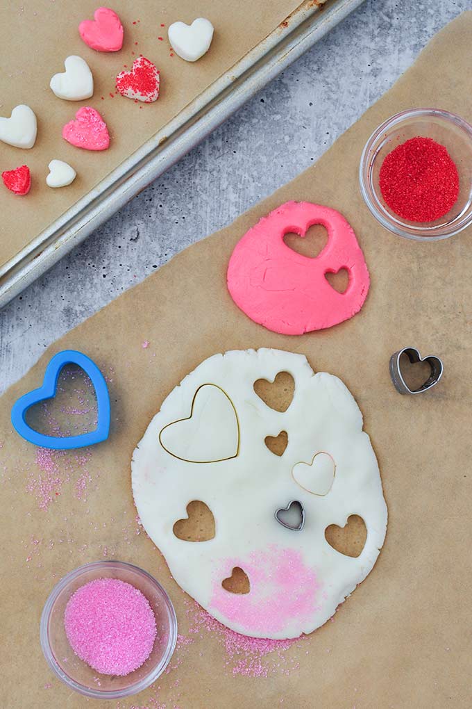 Rolled out cream cheese candy dough with hearts cute out of it, red and pink sugar sprinkles, and heart shaped cookie cutters.