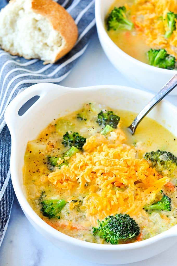 An up close white bowl full of broccoli cheese soup topped with cheese and a bowl of soup and torn off bread in the backround.

