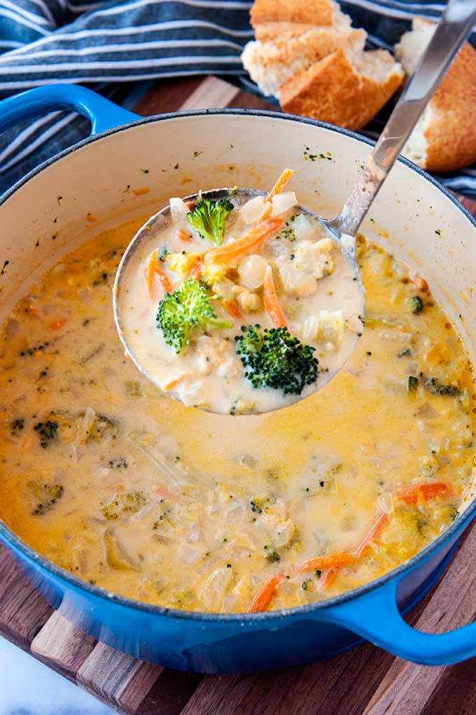 A ladle full of broccoli cheddar soup over a blue pot of soup and torn bread.