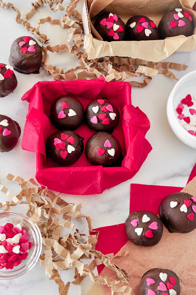 A square box lined with red paper, filled with four truffles.  A rectangular box lined with brown paper with three truffles inside, a small bowl of heart sprinkles, and truffles around both boxes.