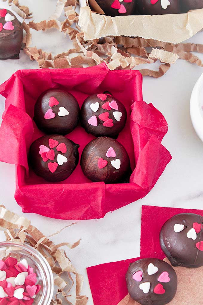 A square box lined with red paper, filled with four truffles.  A rectangular box lined with brown paper with three truffles inside, a small bowl of heart sprinkles, and truffles around both boxes.