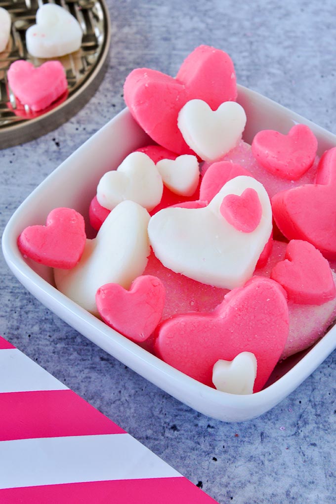 Square bowl of pink and white candy hearts with a striped bag in foreground and a round tray with two small hearts in background.