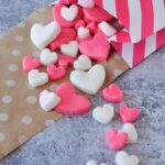 Pink and white Valentine candy hearts in small and big sizes spilling out of a pink and white striped bag onto brown polka dot paper.