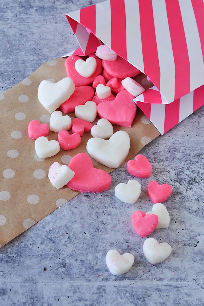 Pink and white Valentine candy hearts in small and big sizes spilling out of a pink and white striped bag onto brown polka dot paper.