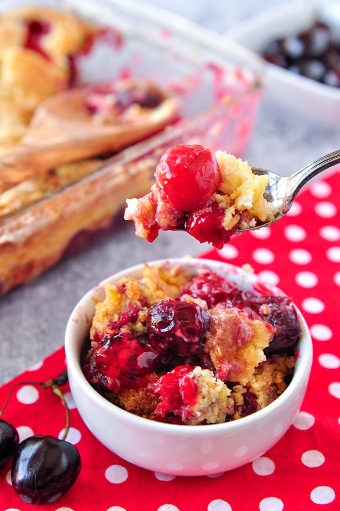 A big spoonful of cherry dump cake above a white bowl and the baking dish in the background.
