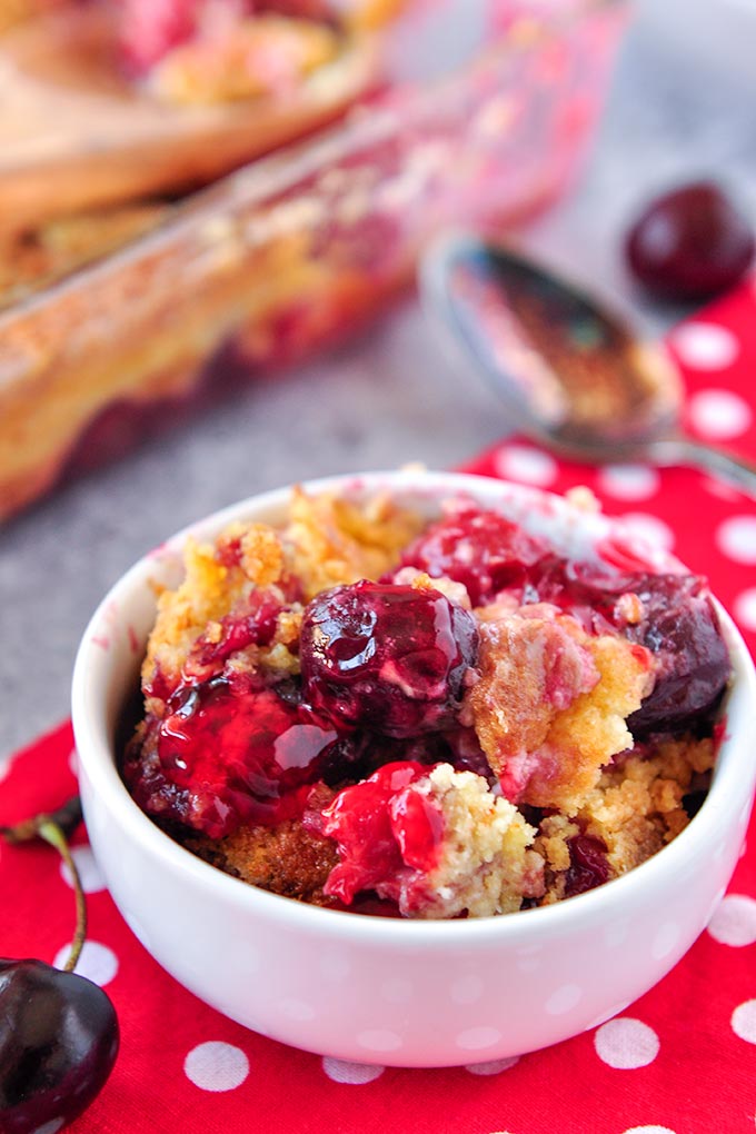 A white bowl full of cherry dump cake with a spoon and a bowl of cherries and more dump cake in the background.