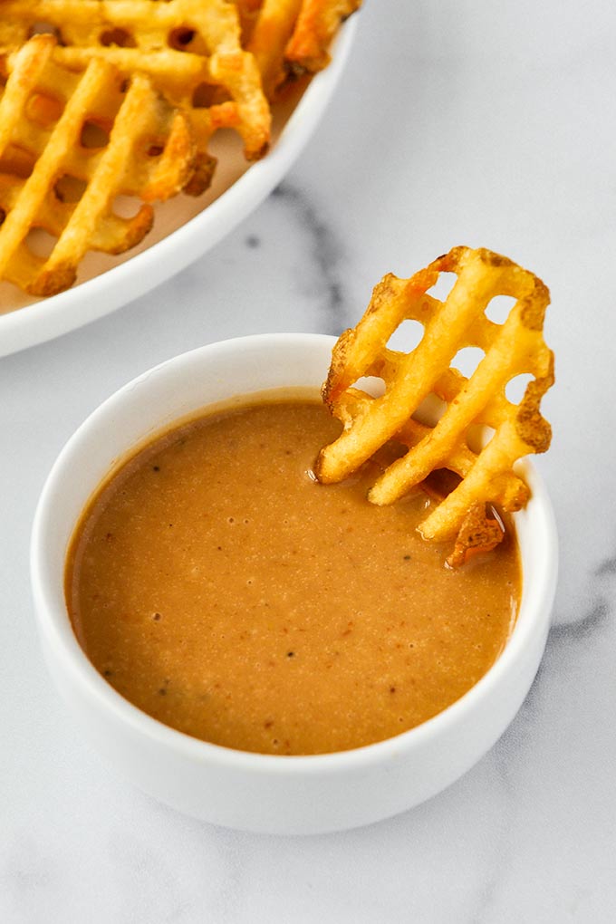 A small white bowl of Chick-fil-A sauce with a waffle fry in it and a plate full of waffle fries in the background.