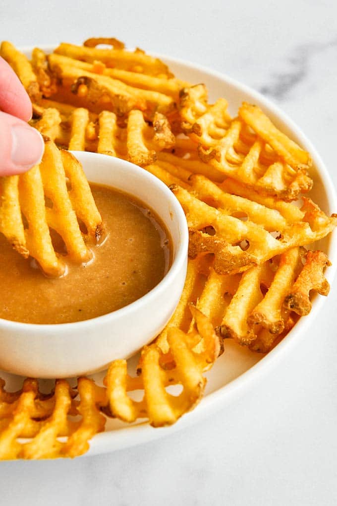 A waffle fry being dipped into a bowl of Chick-fil-A sauce with waffle fries all around.