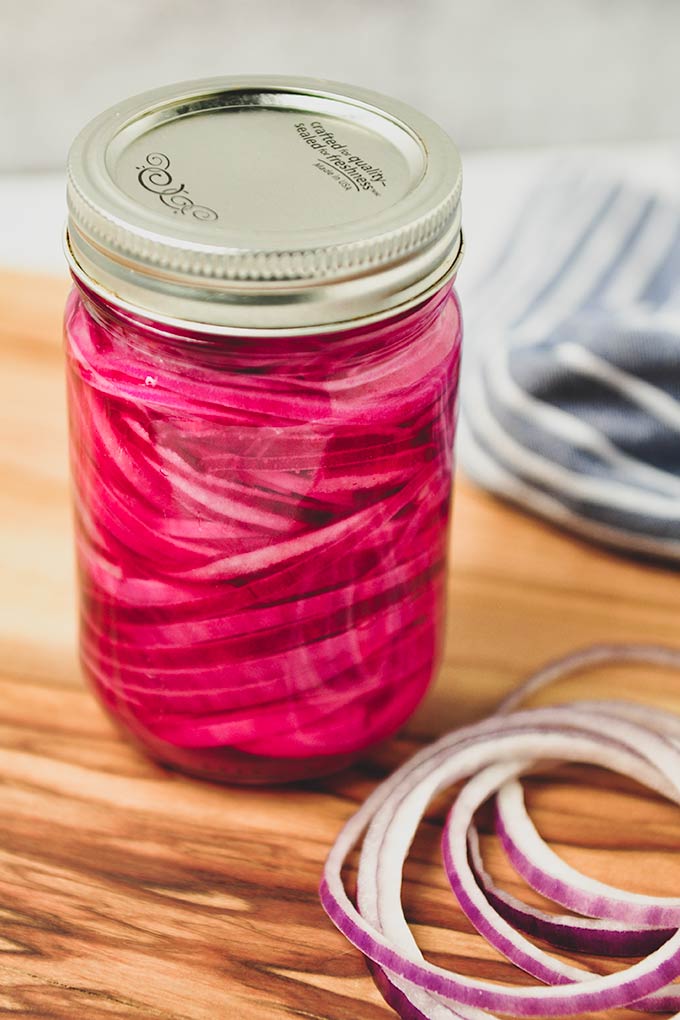 A close up of a large jar with a lid full of red onions with some onions by the jar and a towel