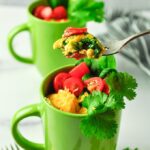 Omelets in two bright green mugs topped with cut up tomatoes, cheese, cilantro and spinach. With a bite of omelet on a fork.