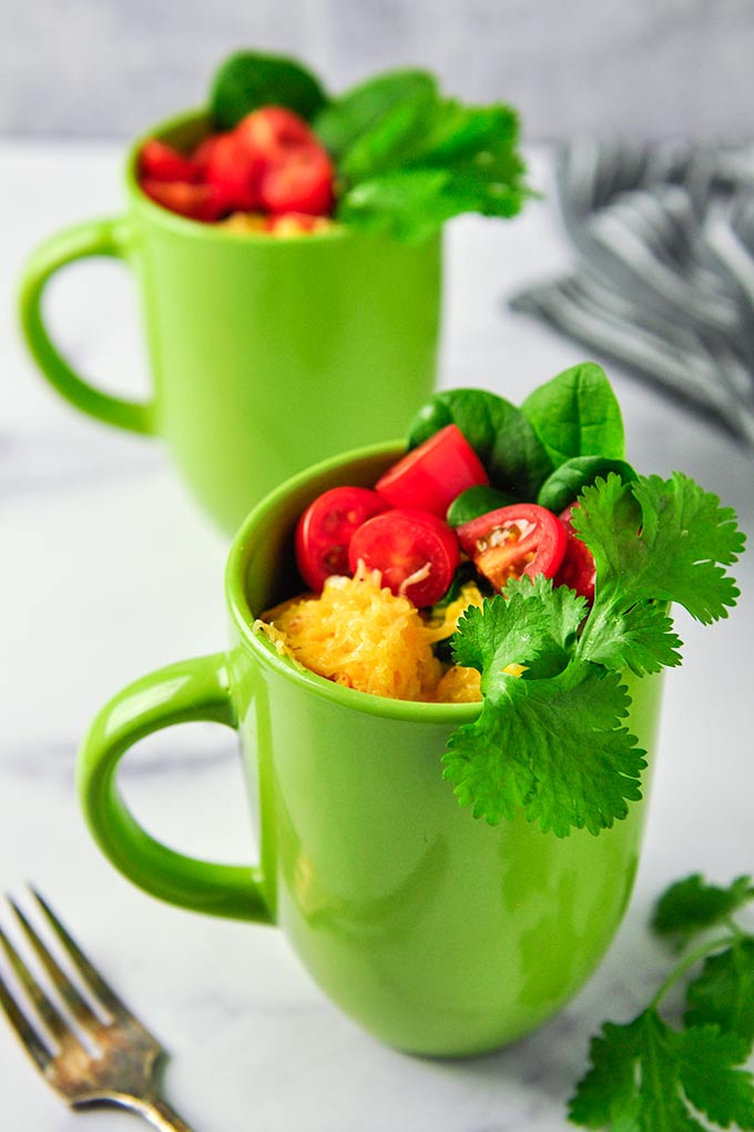 Omelets in two bright green mugs topped with cut up tomatoes, cheese, cilantro and spinach.  