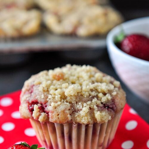 A streusel covered strawberry muffin at the forefront with a strawberry on a red napkin with white polka dots. Bowl of strawberries and muffin tray full of muffins in the background.