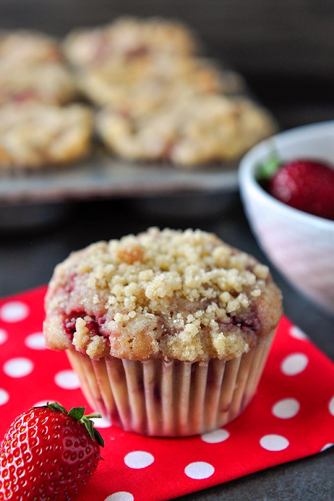 A streusel covered strawberry muffin at the forefront with a strawberry on a red napkin with white polka dots.  Bowl of strawberries and muffin tray full of muffins in the background.