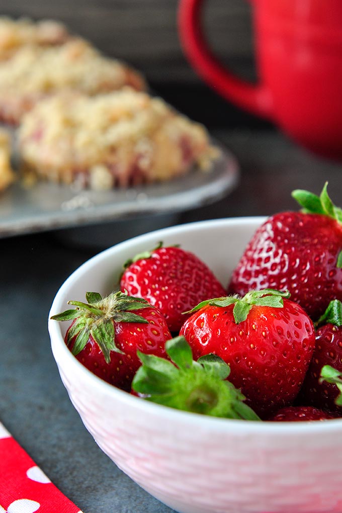 A big bowl of bright red ripe strawberries, tray of muffins and coffee cup in the background.