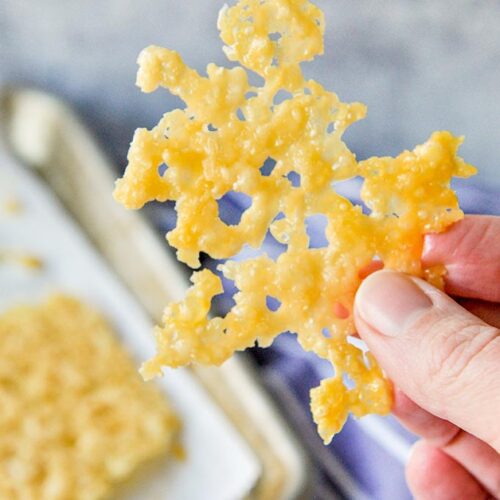 A big piece of a cheese crisp being held. A blue towel and baking sheet of more crisps in the background.