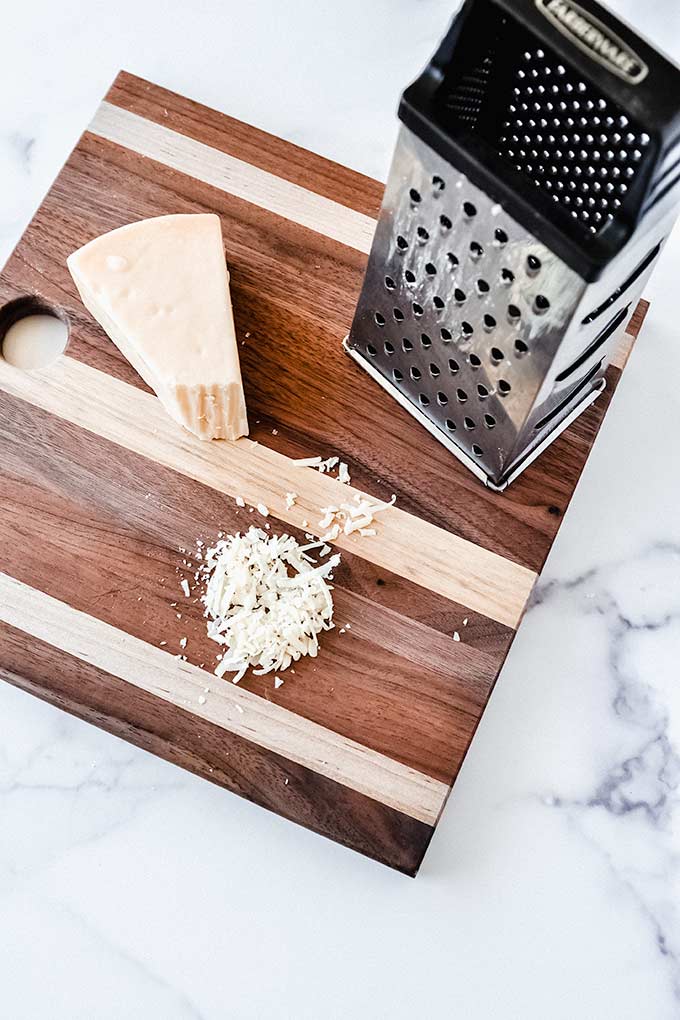 Block of parmesan cheese with a cheese grater on top of a beautiful wood cutting board on a marble background.