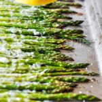 A baking tray with roasted asparagus that is all lined up. A lemon being squeezed over the asparagus is at the top of the image.