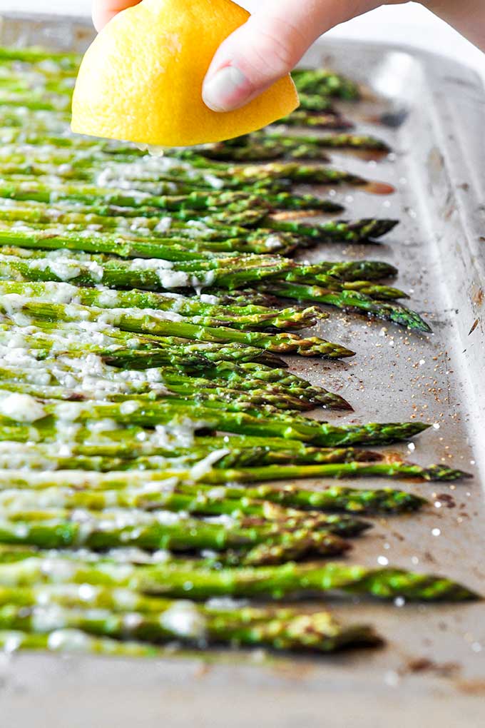 A baking tray with roasted asparagus that is all lined up.  A lemon being squeezed over the asparagus is at the top of the image.