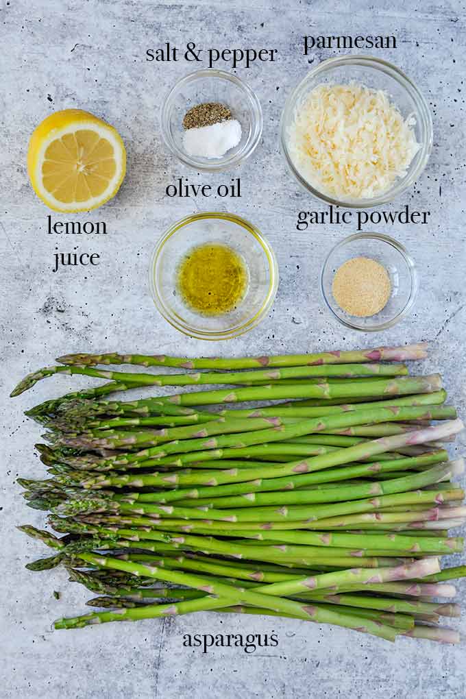 All of the ingredients needed to make baked asparagus such as olive oil, lemon, shredded parmesan cheese, garlic powder and a pound of fresh asparagus.